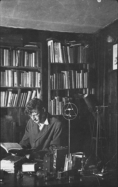 A young man reading a book sits in front of crowded bookcases.