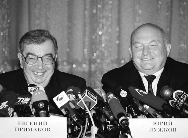 Evgeny Primakov and Iurii Luzhkov smiling and sitting together at a speaker’s platform, August 1999.