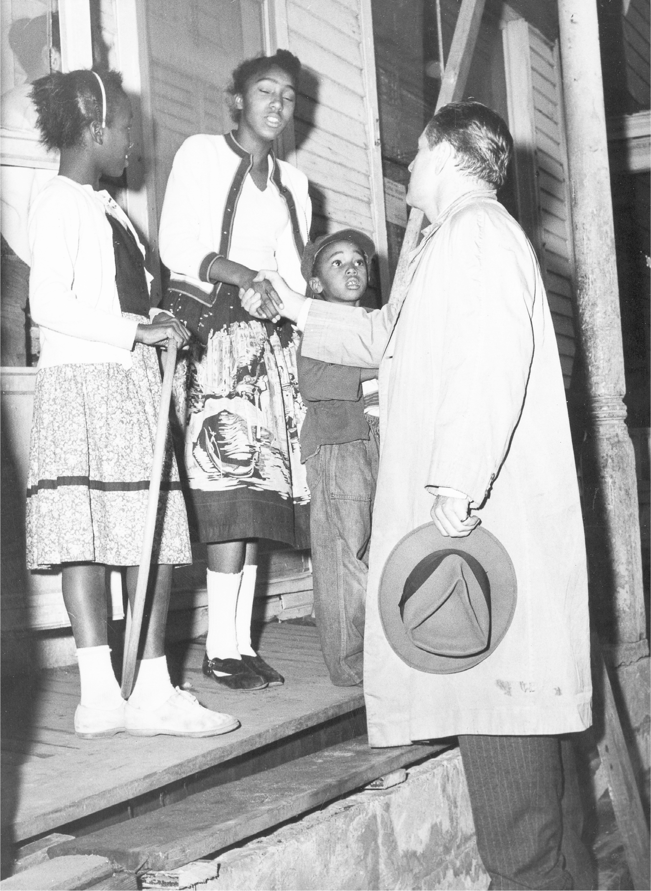 Rockefeller, hat in hand with an overcoat on, greets three children on the front porch of a home. He shakes the hand of the child standing in between the other two children.