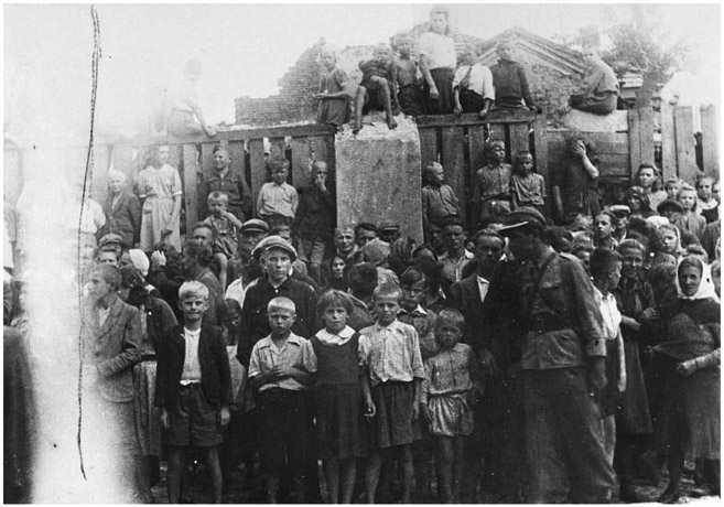 Figure 33. A large group of children stand on the side of a street and sit on fences in the background watching a funeral procession pass by.