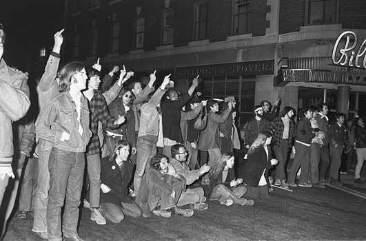 Figure 37. In the dark, a line of young people are taunting police, their middle fingers raised. A few are seated in the street. All are yelling.