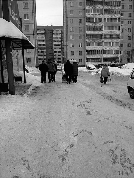 Gray-brown compacted snow covers the ground, with dark patches of ice revealed under some areas. Pedestrians wear puffy winter jackets and hats. Some carry grocery backs. Cars are parked or driving slowly through the parking lot. A grocery store sign and colorful curtains in apartment windows contrast against the beige-gray buildings, snow, and sky.