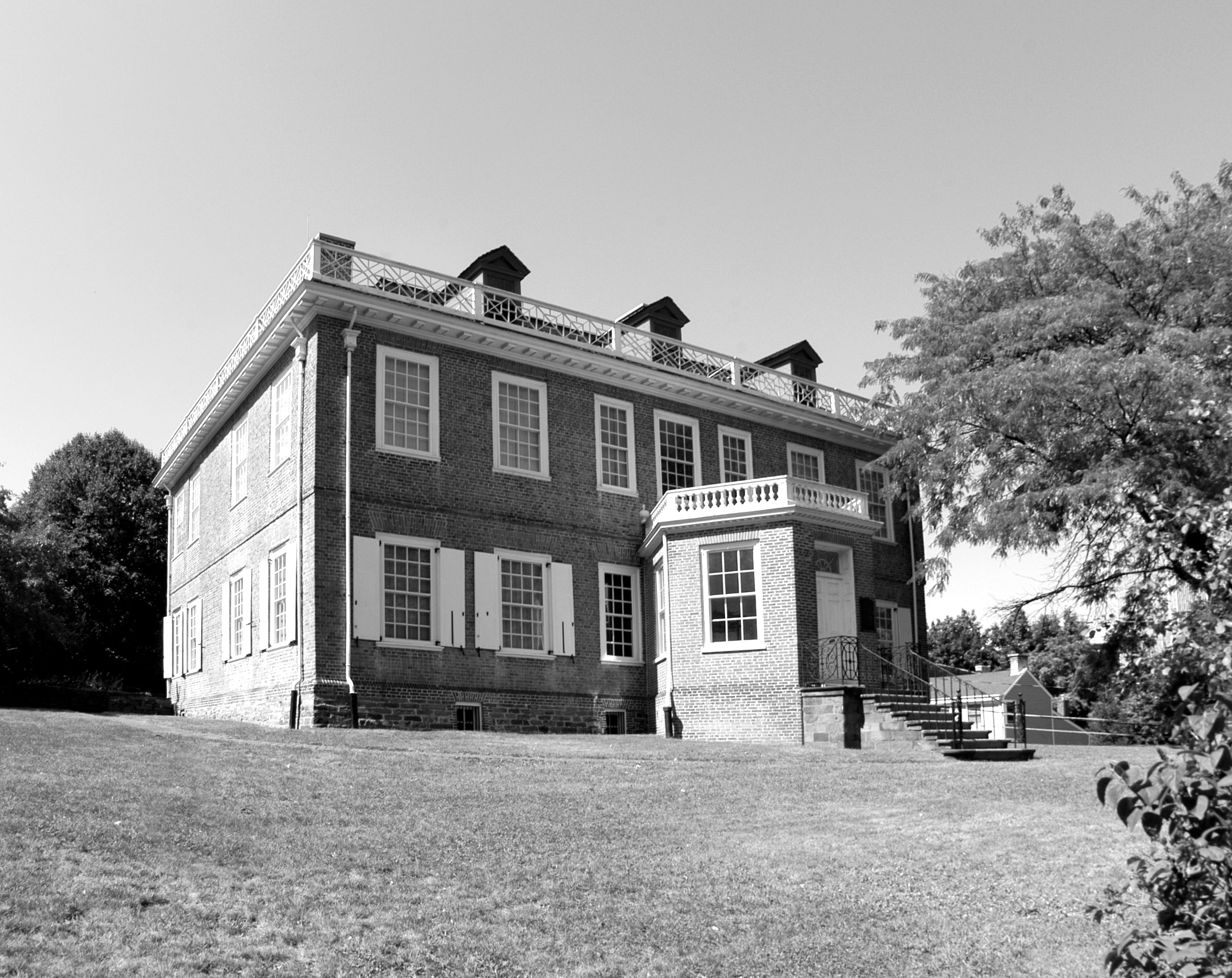 A photograph shows a large Georgian red brick house with open white shutters and a circular vestibule with windows. Steps lead to this main entrance. The house stands on a hill with a lawn and some green trees.