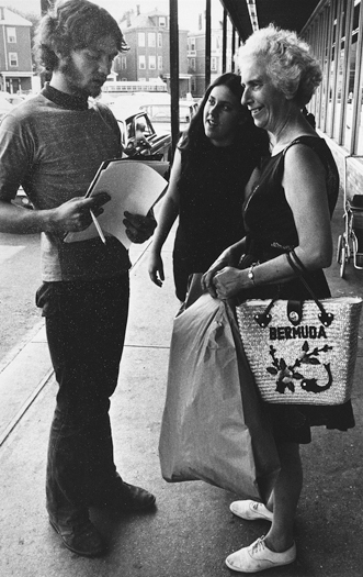 Figure 41. A bearded young man in bell-bottoms is standing talking with two women, one older and one younger outside a store. He holds a petition that he is asking them to sign. He looks serious, and the two women look surprised.