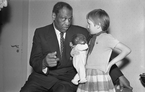 A middle-aged Black man wearing a business suit is in conversation with a young white girl cradling a Black baby doll in her arms.