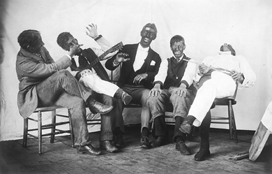 Five white men, seated in chairs and laughing, have their faces smeared with dark pigment and are wearing clownish rustic getups (short pants over trousers, pants with mismatched legs, and baggy workpants).