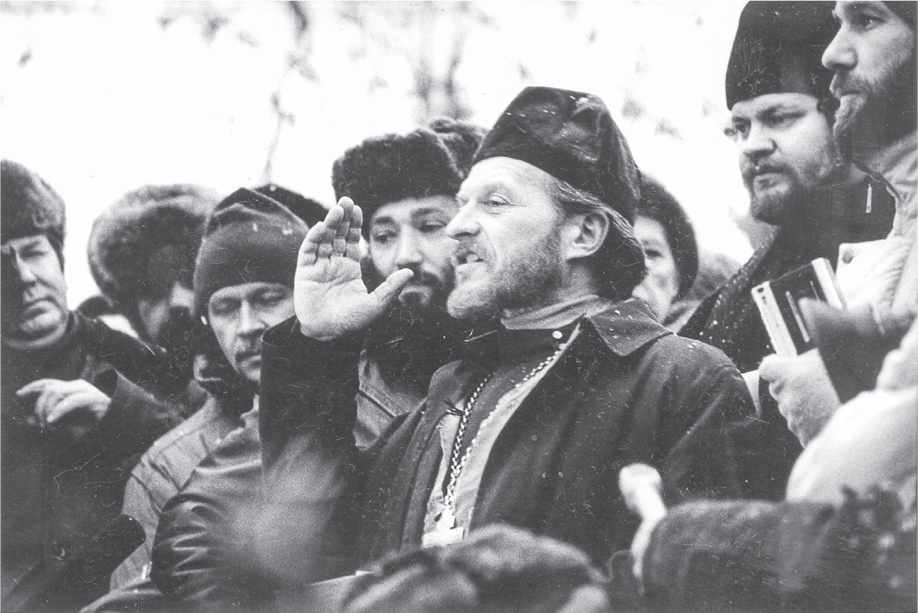 A bearded man wearing a hat and a large cross on a chain speaks to a crowd of men, his right hand gesturing. The men wear winter hats and look thoughtful and serious.