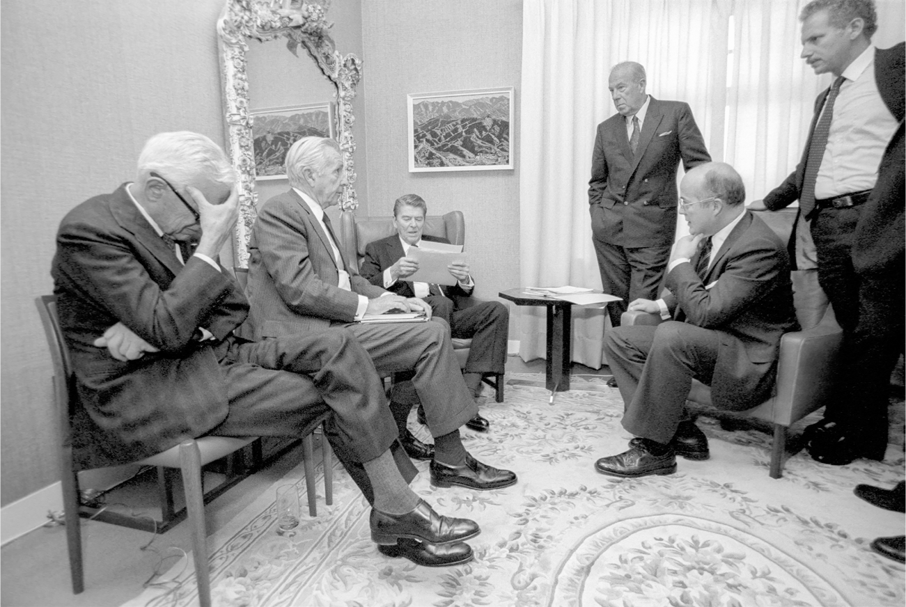 President Reagan sits in a chair in the corner of a small room while his advisers stand or sit to his left and right. Nitze sits in the foreground with his head down and his hand to his face in a look of intense concentration.
