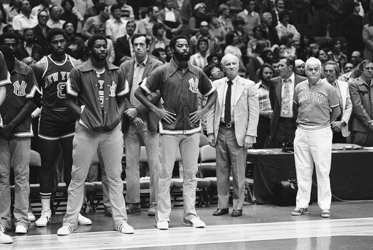 Somber-looking 1976–77 Knicks standing at the sideline with disappointed expressions and slightly defeated postures, such as hands in their pockets, behind their backs, and hanging at their sides. Bill Bradley is out of uniform, in a suit jacket and slacks.