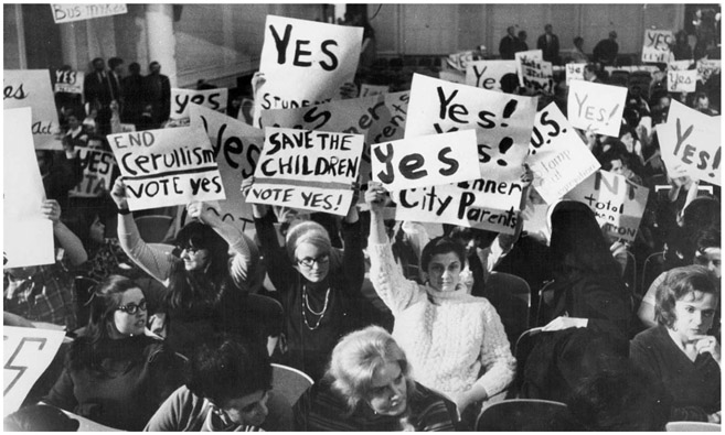 White women sit in rows in a meeting room and hold signs above their heads, including: “Yes!; SAVE THE CHILDREN; END Cerullism VOTE yes.”