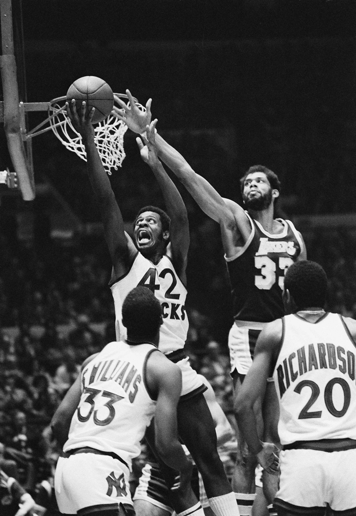 Demic, mouth wide open, jumps up to the hoop for a reverse lay-up as Abdul-Jabbar jumps and reaches from behind, attempting to block the shot.