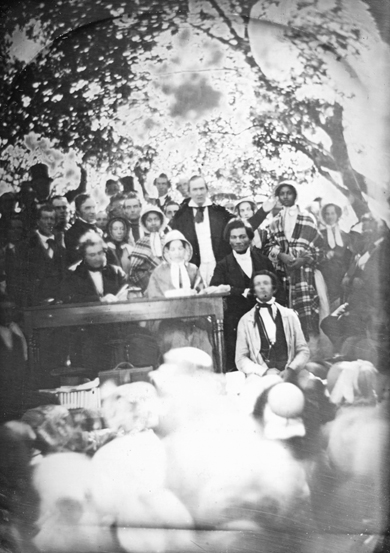 A crowd of fifty men and women, white and Black, pack this fading photograph of conference-goers at an outdoor meeting to protest the passage of the Fugitive Slave Act. The mood is solemn and attentive, and the convention-goers who crowd the lower third of this photograph have their backs to us because they're listening to the presenter.