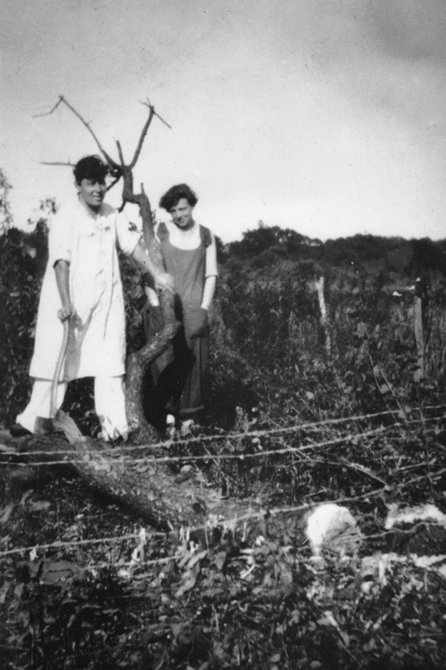 Figure 7. Heap and Anderson, both smiling, stand outside next to a fallen tree. Heap, dressed in a long white smock and pants, places her hand on a branch of the fallen tree, taking a step forward. Anderson, slightly behind Heap, poses with her hands in the front pockets of her overalls.