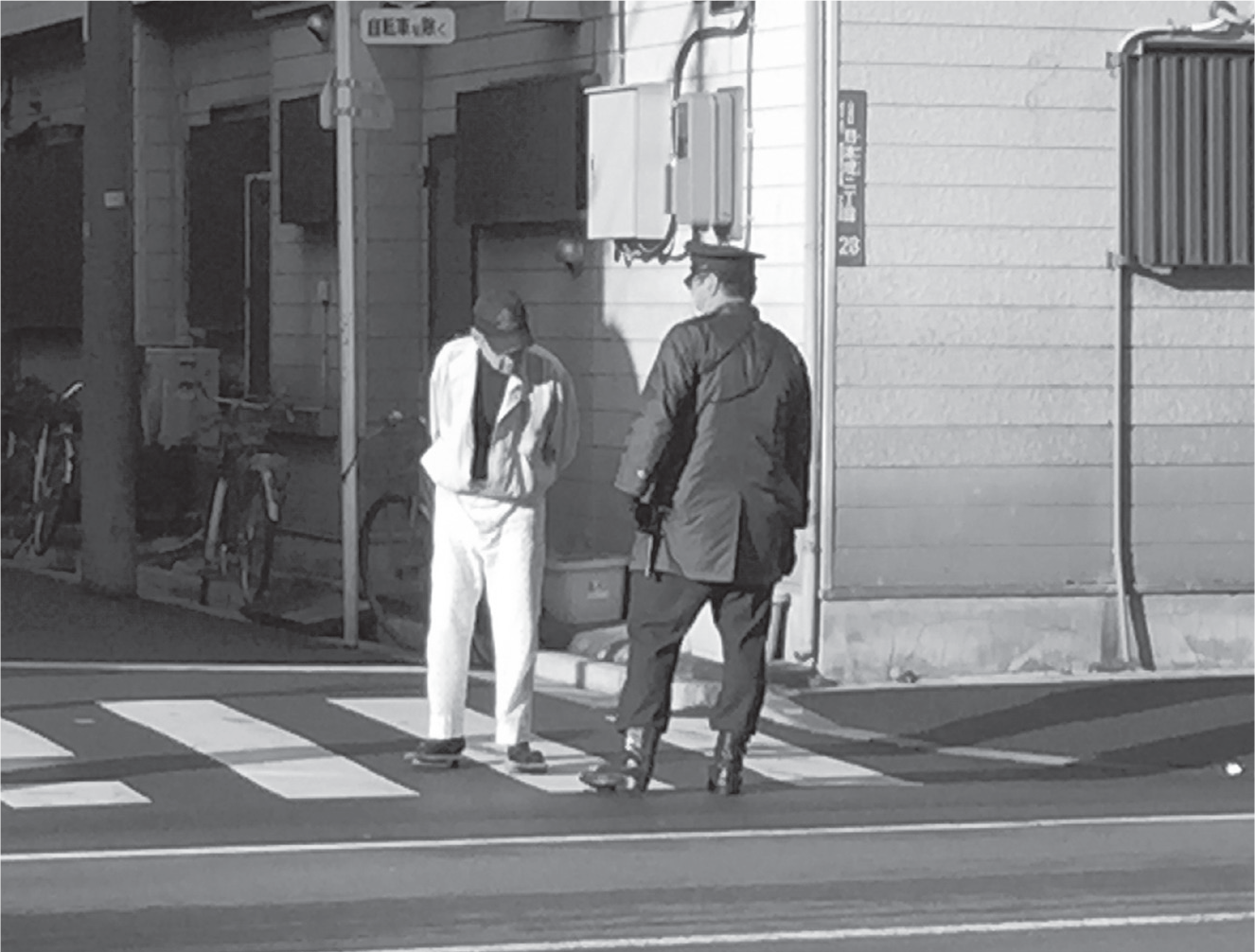 Looking down, an older San’ya resident seems to mutter something as he faces a policeman on the sidewalk. Younger and larger in stature, the policeman holds a baton and rises above the frail figure of the resident.