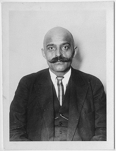 Figure 13. Gurdjieff faces forward with a blank expression, eyes wide and piercing. He wears a dark suit jacket, waistcoat, white shirt and vertical-striped tie. He is bald, with a bushy moustache, waxed at the ends.