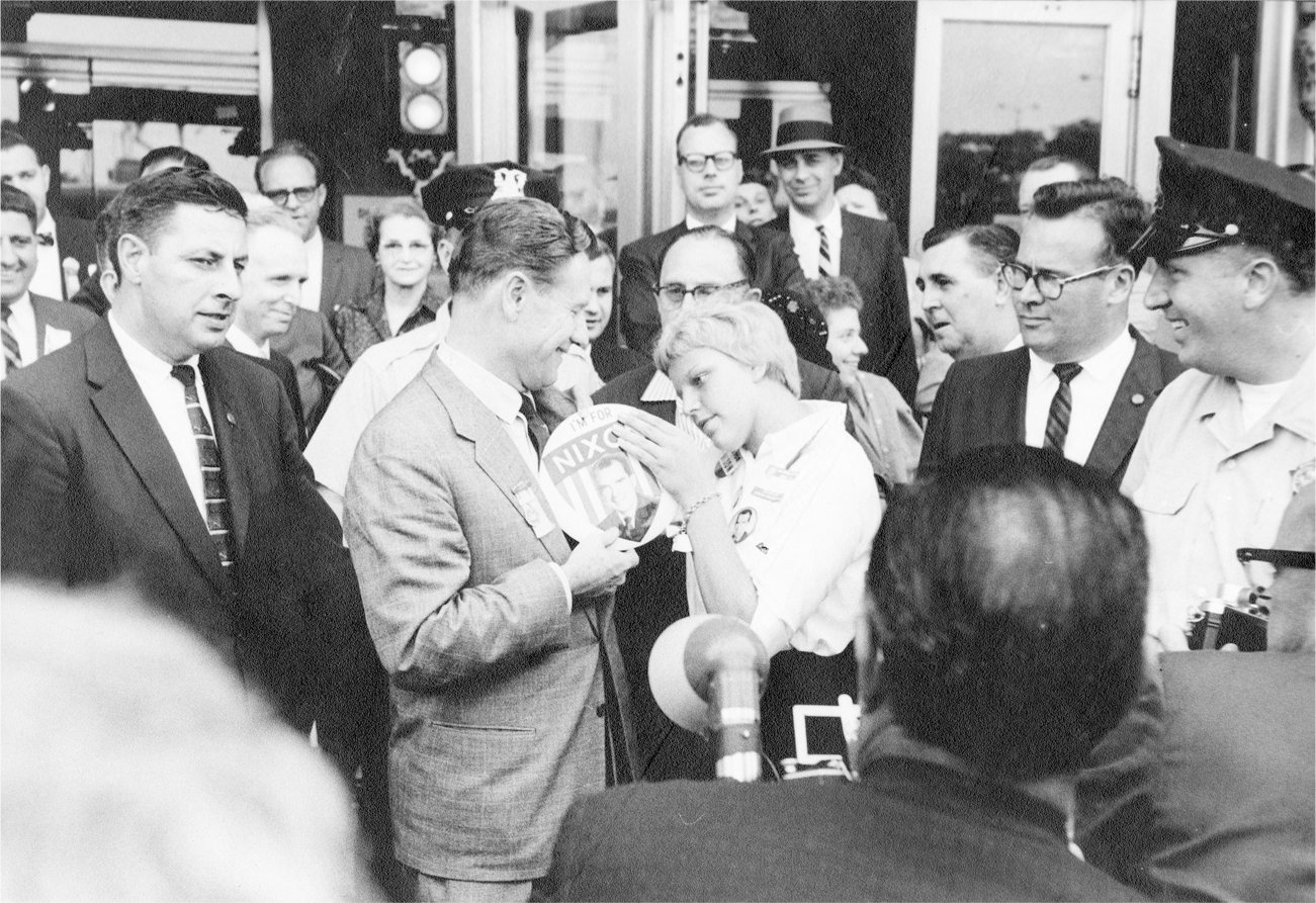 Rockefeller and a teenage girl stand outside at the center of onlookers as she places an oversized pin/button on his lapel. He smiles broadly as she affixes the button, which reads, “I’m for Nixon,” on him.