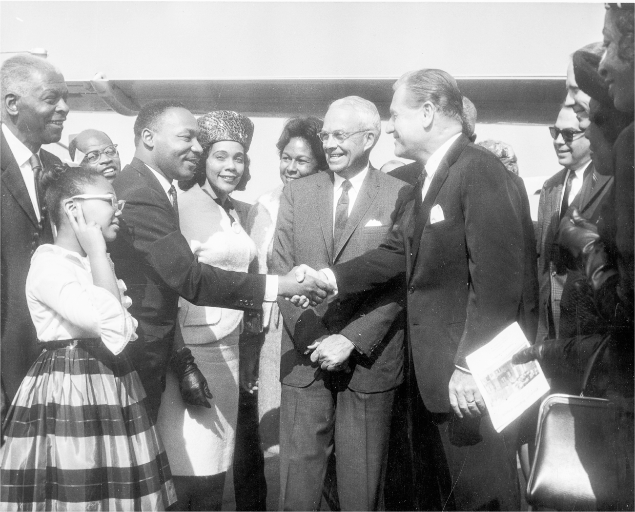 Rockefeller shakes the hand of Martin Luther King Jr., who is flanked by his wife and daughter. Everyone, including the group standing around the pair, are smiling as they greet each other.