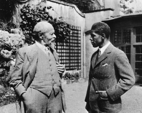 Two men in suits stand outside a cottage, engaged in friendly conversation.