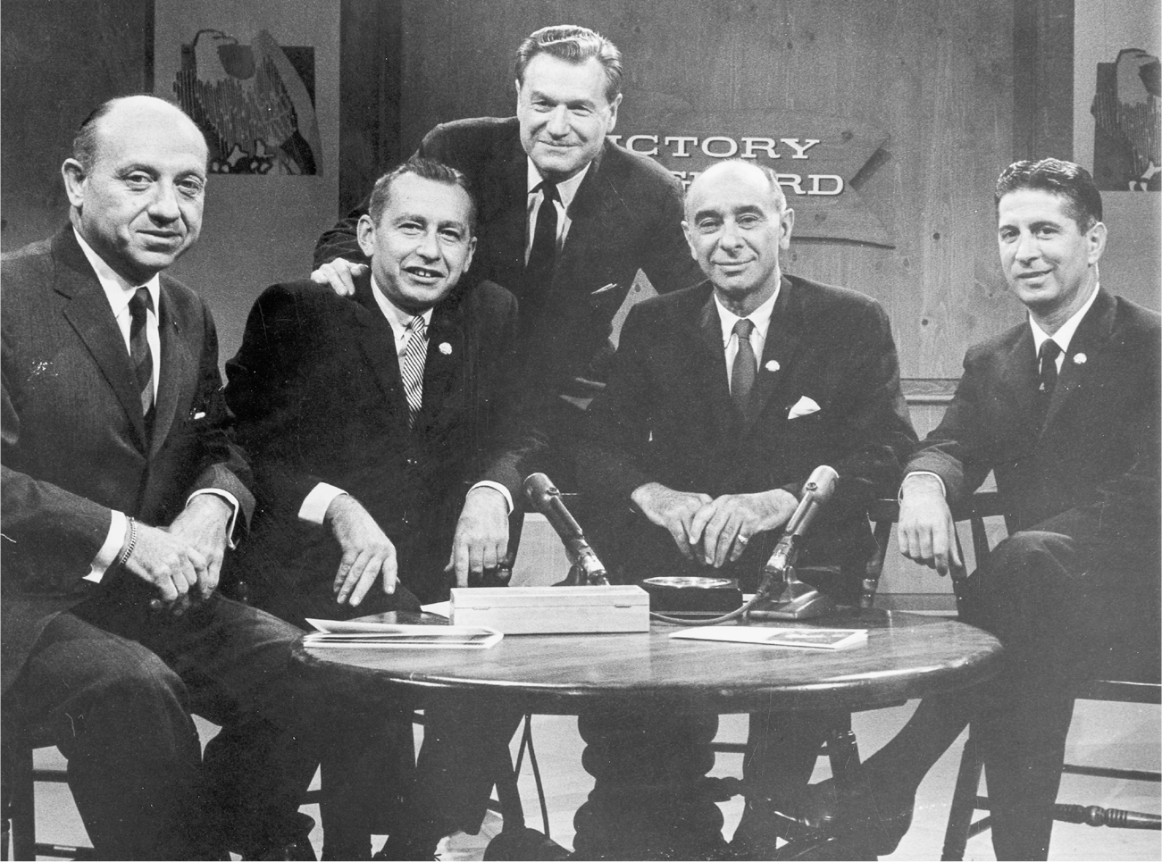 A group of five politicians, including Rockefeller, pose for a photograph. Four of them sit around a table with two microphones on it as Rockefeller leans down from behind to get in the camera frame.