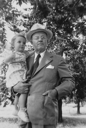 Figure 1 In a summertime scene under a tree, a dapper-looking older man in coat and tie and hat holds a toddler in a floral dress.
