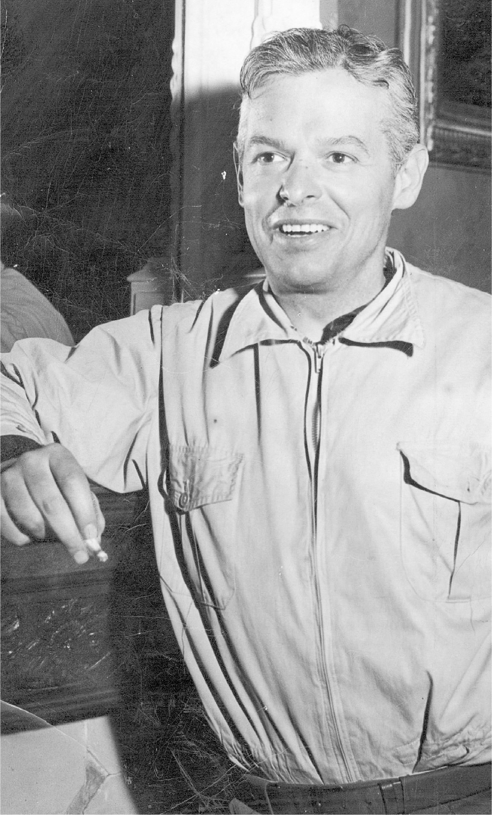A smiling man in his forties looks to the left of the camera and holds a cigarette. The man has short wavy hair and is wearing a light-colored zippered shirt.