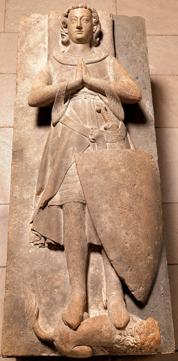 Figure 2. A recumbent knight, head bare, eyes open, hands joined in prayer, wearing chain armor, a sleeveless surcoat, a small belt, a large belt attached to his scabbard from which projects the sword hilt, and ankle straps for spurs. A shield covers his left leg; there is a lion at his feet.