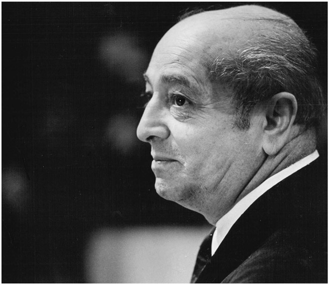 A bald man in a business suit is seen in profile with his lips pursed.