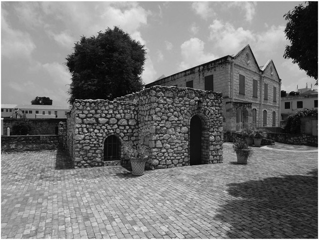 Figure 9.2: A one-story building in the foreground, one part of which is protruding. The walls of the building contain rocks of dissimilar sizes. To the right in the background is a much longer two-story building, the top of whose façade has the shape of a double triangle. In front of both buildings are two flowerpots.