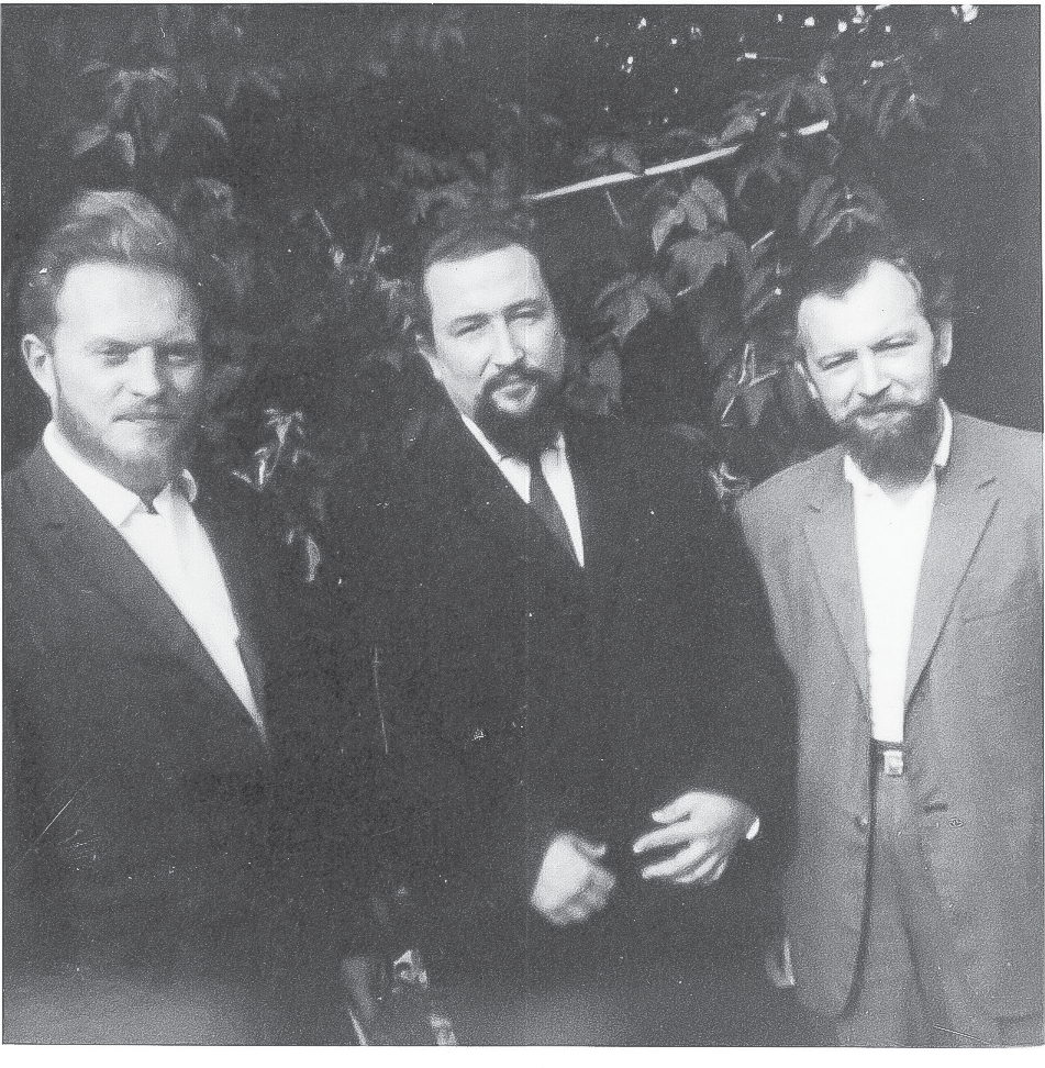 Three bearded men in suit jackets, the center man in a tie, squint slightly in the light. The man on the left has a full head of russet hair, the other two, dark hair.