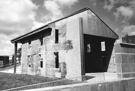 Figure 19 Two parallel plain brick walls stand two stories high, with empty window openings. Minimal steel bracing and a sheltering metal roof have been added. The difference between the original bricks and those added later is clear.