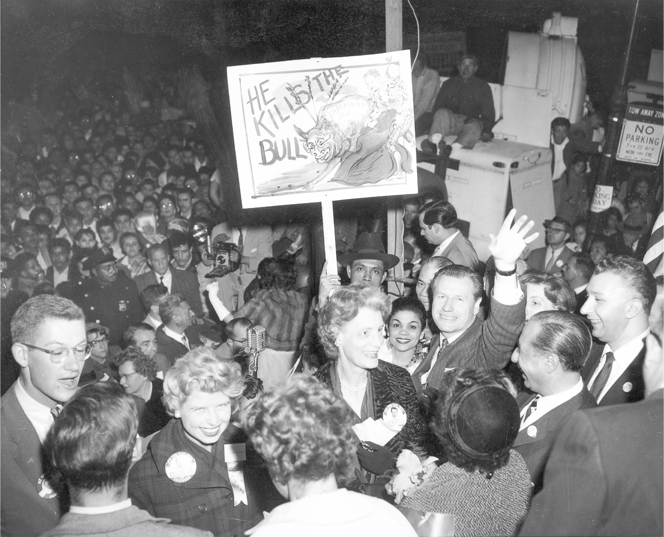 Rockefeller waves at the camera with a big smile while standing in a huge crowd of people. A spectator behind him holds a poster that depicts a drawing of Rockefeller as a matador subduing a bull that is intended to represent Tammany Hall. The sign reads, “He kills the bull.”