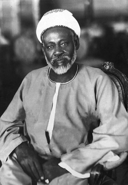 Photograph of a man with a graying beard sitting in a chair. He is wearing a robe and a headdress. He looks directly at the camera with his hands in his lap.