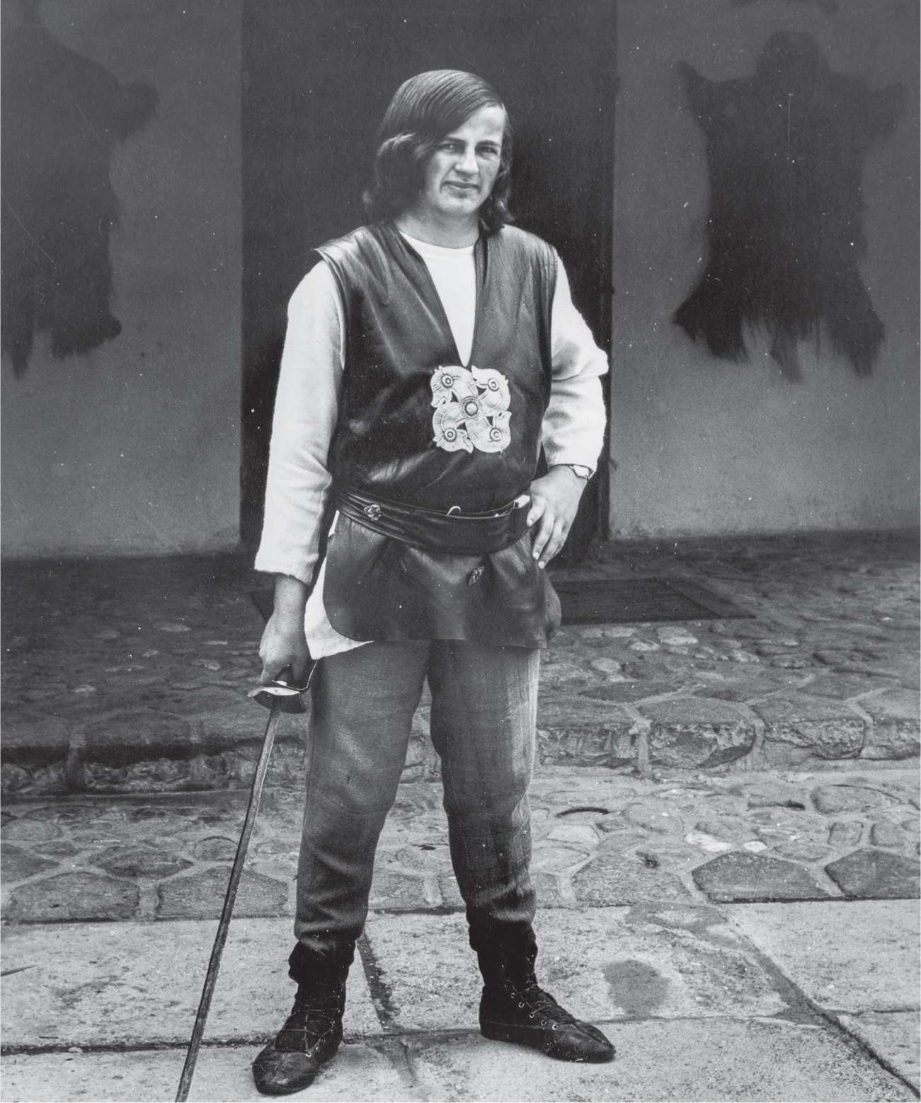 A man with long hair wears a traditional costume and holds a sword. He is standing in front of a building with animal skins on its walls.