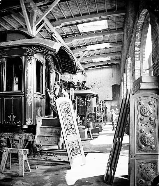 Factory worker painting the doorframe of a railroad car with five ornate doors nearby. On the ornate door in the foreground is visible the letters PPCCo.