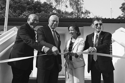 Three men and one woman stand holding a ribbon in front of a building. The men are all in dark suits, while the woman is wearing a light-colored suit. The men look to the right of the image, while the woman looks to the left.