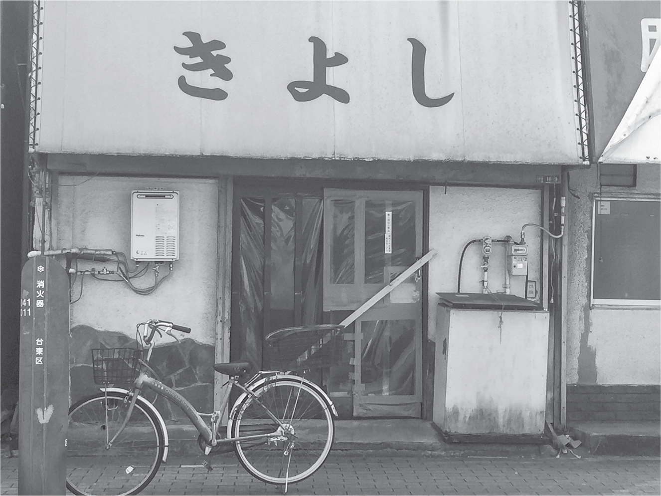 Placed diagonally, a wooden board holds the sliding doors of a dive bar in place. The windows of the sliding door have been replaced with plastic sheets, and a bicycle stands in front of the musty, faded storefront.