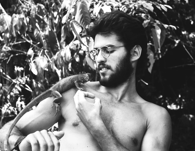 A green iguana is sitting on Marcelo’s shoulder, and the two of them are looking at each other.