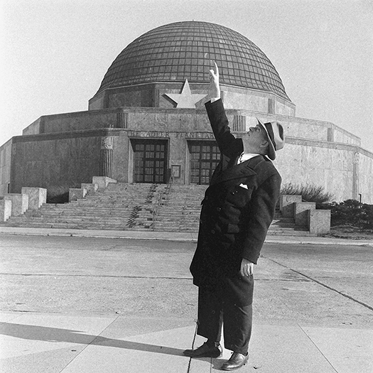 A man wearing a suit and a hat stands in front of a domed building and points at the sky.