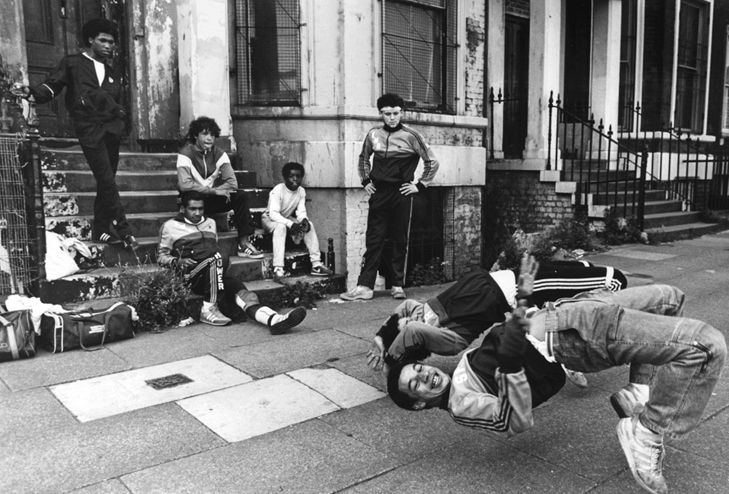 Two members of the Eastwood Rockers breakdance on a sidewalk, while five older kids watch from the front stoop of an apartment building.