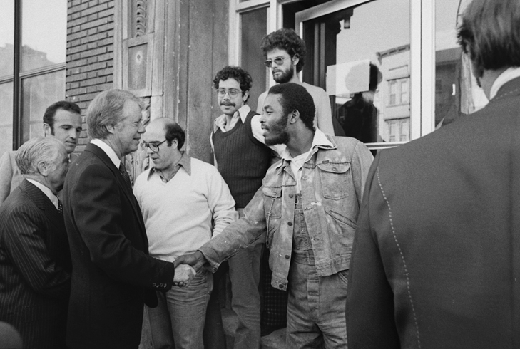 President Jimmy Carter shakes hands with a young Black man in front of a South Bronx apartment building while others look on.