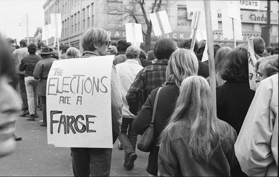 Figure 19. A long line of protesting young people is seen from the back, marching on election day. Many are carrying signs. One young man has his sign on his back so you can see that it reads “The elections are a farce.”