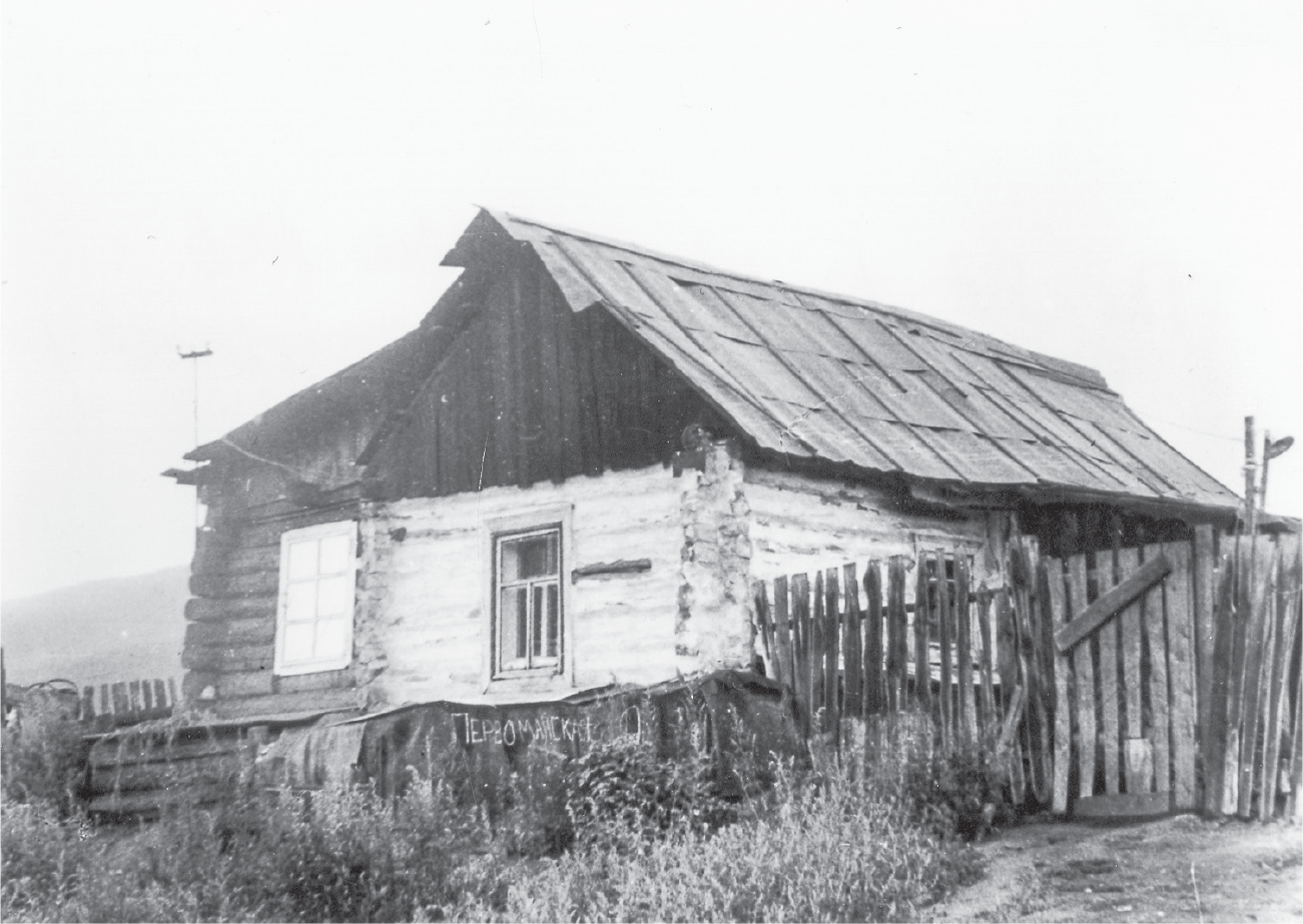 A weathered log cabin with a tin roof and three windows, one of them (left) covered, and a rough wooden fence on the right.