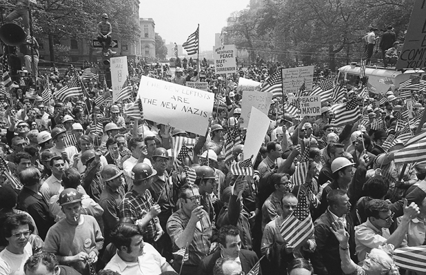 Figure 28. Young and middle-aged white men, many in hard hats, waving American flags, crowd onto a New York street. One is sitting on the top of a one-way street sign. A placard says, “The new left are the new Nazis.” Another says” Win a negotiated peace but no surrender.”