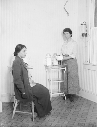Figure 7.5. Margaret Sanger and her assistant, Fania Mindell, are photographed in a small, spare room in the Brownsville clinic. Sanger sits in a wooden chair, staring blankly forward. Mindell stands next to a small table with a bowl in her hands. There is nothing else in the room.