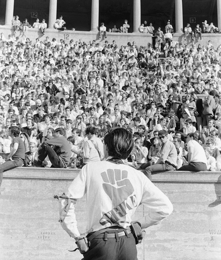 Figure 23. Thousands of students sit on rows of stadium seats with the spring sun beating down. A student strike marshal stands with his back to the camera, one hand on his hip, wearing a t-shirt with a large red fist and the word “STRIKE.”