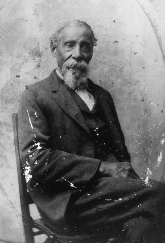 Dressed semiformally and seated in a plain wood chair, an older man with a jaunty Vandyke beard gazes pensively at the camera.