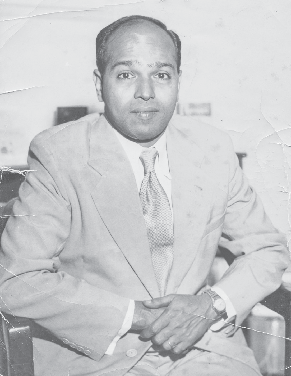 Uncle Mohan, dressed in a light-colored suit and tie, sits in a chair in his office with his hands clasped together in front, his wedding ring and watch in full view, as he looks straight at the camera.