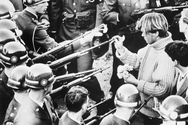 Figure 13. A young man wearing a bulky turtleneck sweater holds flowers in one hand, while with the other he places a flower in the barrel of a rifle held by a soldier.