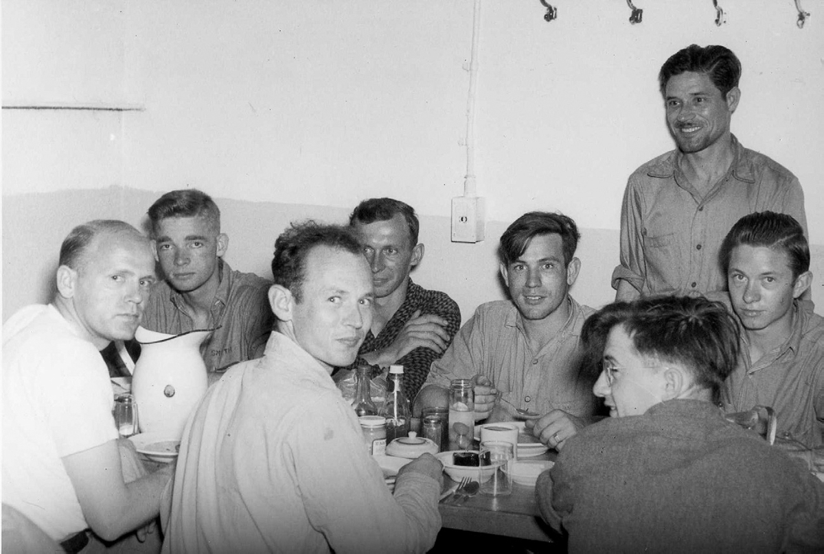 Figure 3. Seven men sit around a table of food, while an eighth stands behind them. The three men with their backs to the camera have their heads turned to look at the photographer.