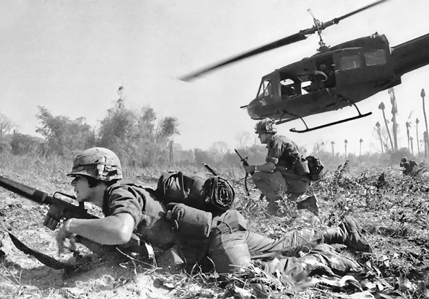 Figure 7. A soldier lies on the ground; another kneels. Above them one attack helicopter hovers. All around them is debris of trees and plants pulverized by bombings.